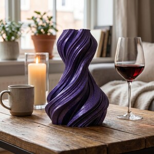 K&ouml;nnte beinhalten: Eine dekorative, wirbelnde lila Vase mit glitzerndem Finish steht auf einem Holztisch. Eine brennende Kerze in einem Glashalter, eine Kaffeetasse und ein Weinglas mit Rotwein stehen ebenfalls auf dem Tisch. Die Vase hat ein einzigartiges, strukturiertes Design.