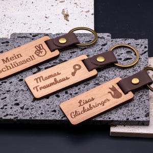 rei personalisierte Schlüsselanhänger aus Holz mit Kunstlederband und Messingring, graviert mit individuellen Texten: „Mein Schlüssel!“ mit Peace-Handsymbol, „Mamas Traumhaus“ mit Schlüssel-Symbol und „Lisas Glücksbringer“ mit Katzenmotiv. Die rechteckigen Anhänger liegen auf strukturierten Steinplatten und kombinieren rustikale Natürlichkeit mit modernem Design. Ideal als Geschenkidee für Familie oder Freunde, praktisch, dekorativ und langlebig.