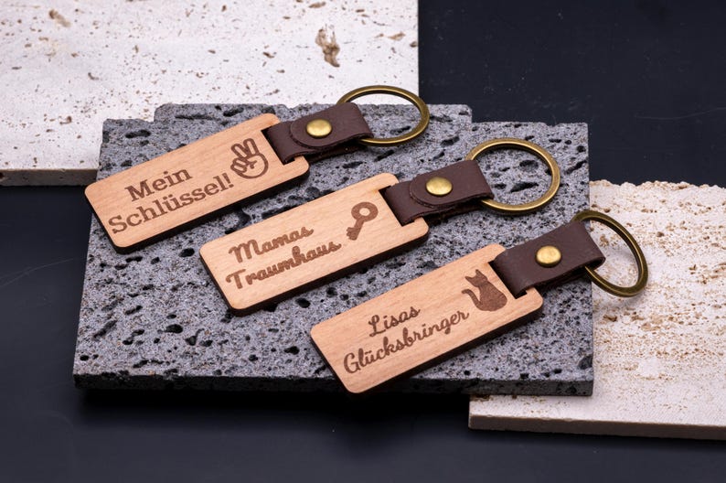 rei personalisierte Schlüsselanhänger aus Holz mit Kunstlederband und Messingring, graviert mit individuellen Texten: „Mein Schlüssel!“ mit Peace-Handsymbol, „Mamas Traumhaus“ mit Schlüssel-Symbol und „Lisas Glücksbringer“ mit Katzenmotiv. Die rechteckigen Anhänger liegen auf strukturierten Steinplatten und kombinieren rustikale Natürlichkeit mit modernem Design. Ideal als Geschenkidee für Familie oder Freunde, praktisch, dekorativ und langlebig.