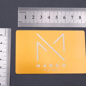 Hellgoldene Metall-Visitenkarte aus Aluminium mit modernem Mango-Digitals-Logo in matter Gravur. Daneben liegen Lineale, die die Größe der Karte verdeutlichen (ca. 8,5 x 5,4 cm). Die stabile Karte mit abgerundeten Kanten hebt sich durch ihre exklusive Materialwahl von klassischen Papierkarten ab und eignet sich perfekt als elegante Businesskarte für Firmen mit hohem Anspruch an Design und Langlebigkeit.