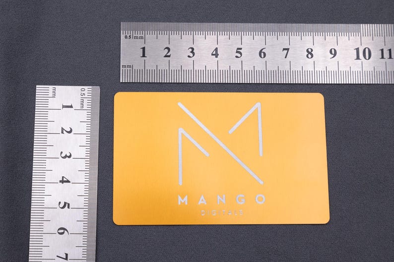 Hellgoldene Metall-Visitenkarte aus Aluminium mit modernem Mango-Digitals-Logo in matter Gravur. Daneben liegen Lineale, die die Größe der Karte verdeutlichen (ca. 8,5 x 5,4 cm). Die stabile Karte mit abgerundeten Kanten hebt sich durch ihre exklusive Materialwahl von klassischen Papierkarten ab und eignet sich perfekt als elegante Businesskarte für Firmen mit hohem Anspruch an Design und Langlebigkeit.