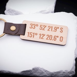 Ein gravierter Holz Schlüsselanhänger mit Koordinaten „33° 52' 21.9'' S, 151° 12' 20.6'' O“ liegt auf weißem Stein. Helles Holz, kombiniert mit braunem Lederband und stabilem Metallring in Antik-Gold, sorgt für einen edlen Look. Der Anhänger ist ideal für eine personalisierte Gravur mit Koordinaten, Namen oder Sprüchen. Ein besonderes Geschenk, das Erinnerungen an Orte festhält und stilvoll am Schlüsselbund getragen werden kann.