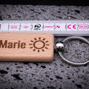 Rechteckiger Schlüsselanhänger aus Buchenholz, gemessen ca. 5 cm Länge mit stabilem Metallring, graviert mit dem Namen Marie und einem Sonnensymbol. Das Maßband zeigt die exakte Größe des Anhängers und unterstreicht die hochwertige Verarbeitung. Perfekt als personalisiertes Geschenk mit langlebiger Lasergravur, ideal für Schlüsselbund, Tasche oder Rucksack. Ein praktisches, stilvolles und individuelles Accessoire für jeden Anlass.