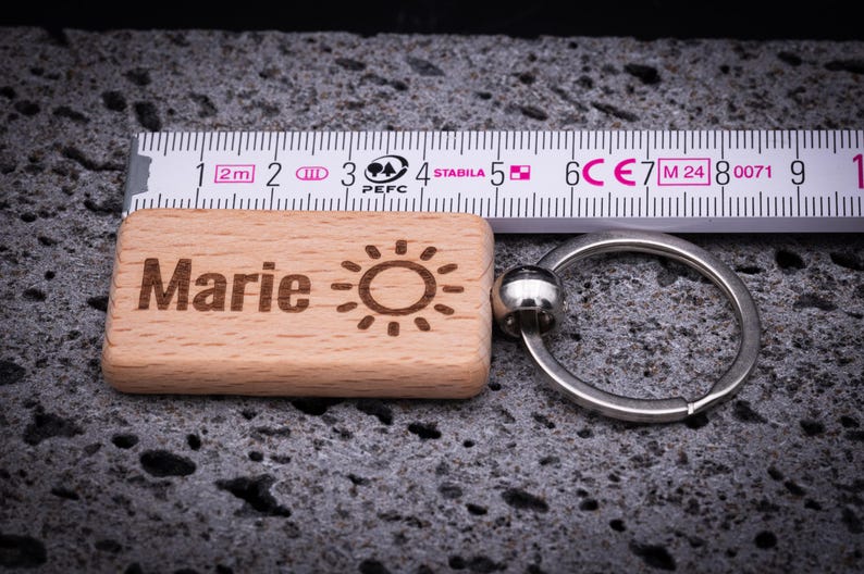 Rechteckiger Schlüsselanhänger aus Buchenholz, gemessen ca. 5 cm Länge mit stabilem Metallring, graviert mit dem Namen Marie und einem Sonnensymbol. Das Maßband zeigt die exakte Größe des Anhängers und unterstreicht die hochwertige Verarbeitung. Perfekt als personalisiertes Geschenk mit langlebiger Lasergravur, ideal für Schlüsselbund, Tasche oder Rucksack. Ein praktisches, stilvolles und individuelles Accessoire für jeden Anlass.