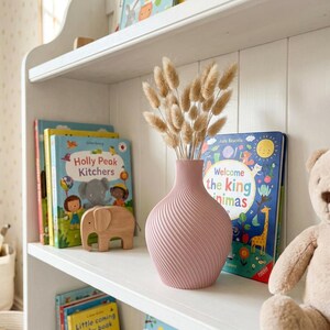 Puede incluir: Un jarr&oacute;n rosa con dise&ntilde;o en espiral contiene flores secas, colocado en una estanter&iacute;a blanca. Libros infantiles con cubiertas coloridas y una figura de elefante de madera tambi&eacute;n est&aacute;n en el estante. Un oso de peluche est&aacute; cerca.