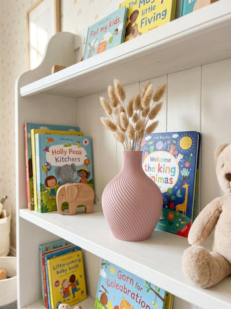 Puede incluir: Un jarr&oacute;n rosa con dise&ntilde;o en espiral contiene flores secas, colocado en una estanter&iacute;a blanca. Libros infantiles con cubiertas coloridas y una figura de elefante de madera tambi&eacute;n est&aacute;n en el estante. Un oso de peluche est&aacute; cerca.