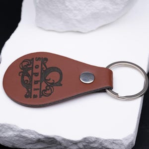 Liegend präsentierter personalisierter Schlüsselanhänger aus braunem Kunstleder mit eingraviertem Namen „Sophia“. Die aufwendige Monogrammgestaltung mit floralen Linien und klarer Typografie unterstreicht den edlen Look des Accessoires. Dank stabilem Metallring und sauberer Verarbeitung ist der Anhänger langlebig und alltagstauglich. Eine stilvolle Geschenkidee für Männer und Frauen, die Wert auf Individualität legen.