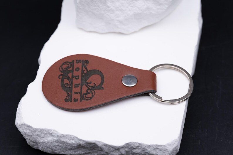 Liegend präsentierter personalisierter Schlüsselanhänger aus braunem Kunstleder mit eingraviertem Namen „Sophia“. Die aufwendige Monogrammgestaltung mit floralen Linien und klarer Typografie unterstreicht den edlen Look des Accessoires. Dank stabilem Metallring und sauberer Verarbeitung ist der Anhänger langlebig und alltagstauglich. Eine stilvolle Geschenkidee für Männer und Frauen, die Wert auf Individualität legen.