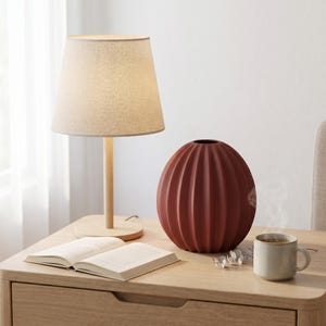 May include: A beige lampshade on a wooden lamp base, a terracotta-coloured vase, a mug, and an open book rest on a wooden bedside table. The vase has vertical ridges. A cup of coffee sits next to the vase.