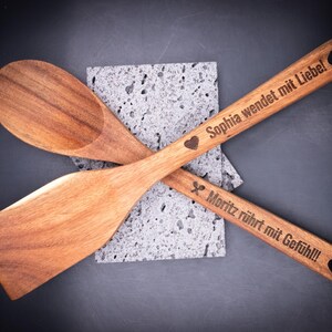 Zwei gravierte Holzlöffel mit Gravurtexten „Sophia wendet mit Liebe!“ und „Moritz rührt mit Gefühl!!!“ auf dunklem Hintergrund. Das Laserdesign zeigt Symbole und persönliche Texte. Ideal für personalisierte Geschenkideen – langlebig, dekorativ und aus hochwertigem Akazienholz gefertigt.