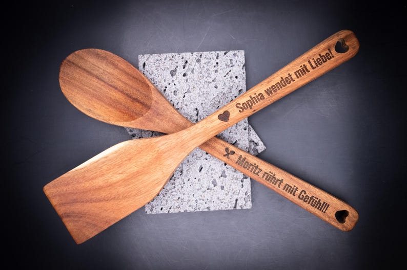 Zwei gravierte Holzlöffel mit Gravurtexten „Sophia wendet mit Liebe!“ und „Moritz rührt mit Gefühl!!!“ auf dunklem Hintergrund. Das Laserdesign zeigt Symbole und persönliche Texte. Ideal für personalisierte Geschenkideen – langlebig, dekorativ und aus hochwertigem Akazienholz gefertigt.