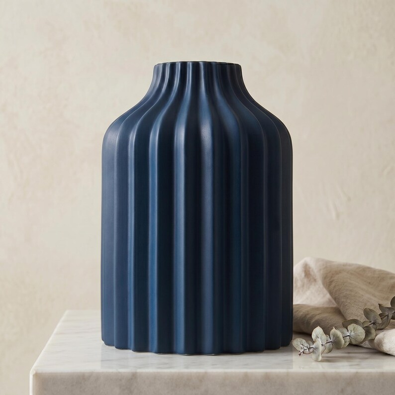 Może przedstawiać: Granatowy ceramiczny wazon z pionowymi żebrowanymi detalami. Wazon ma cylindryczny kształt z lekko rozszerzoną g&oacute;rą. Stoi na białej marmurowej powierzchni. Beżowa szmatka i gałązka zieleni znajdują się w tle.