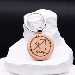 Ein einzelner runder Schlüsselanhänger aus Holz mit Gravur des Sternzeichens Schütze steht auf weißem Steinpodest. Graviert ist das astrologische Symbol des Bogenschützen, eingerahmt von kleinen Sternen, Mond und Schriftzug „Schütze“. Der silberne Schlüsselring ergänzt das natürliche Holzdesign. Die Gravur ist präzise ausgeführt und macht den Schlüsselanhänger zu einem eleganten und personalisierbaren Accessoire für Sternzeichen-Liebhaber.