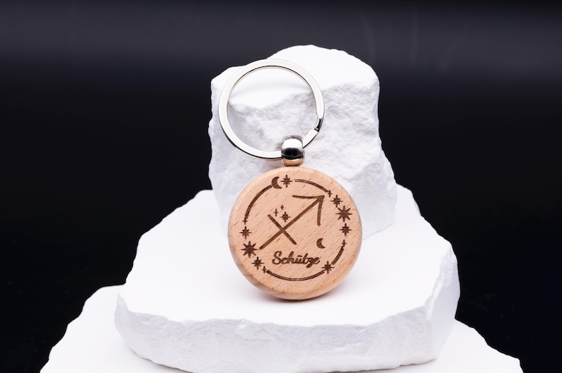 Ein einzelner runder Schlüsselanhänger aus Holz mit Gravur des Sternzeichens Schütze steht auf weißem Steinpodest. Graviert ist das astrologische Symbol des Bogenschützen, eingerahmt von kleinen Sternen, Mond und Schriftzug „Schütze“. Der silberne Schlüsselring ergänzt das natürliche Holzdesign. Die Gravur ist präzise ausgeführt und macht den Schlüsselanhänger zu einem eleganten und personalisierbaren Accessoire für Sternzeichen-Liebhaber.