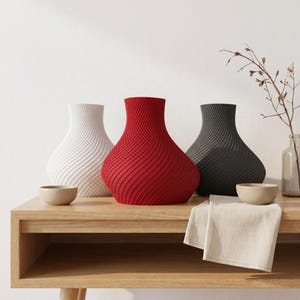 May include: Three decorative vases in white, red, and gray, with a textured, woven design. The vases are displayed on a light wood table, alongside small bowls and a folded linen cloth. A minimalist home decor theme.