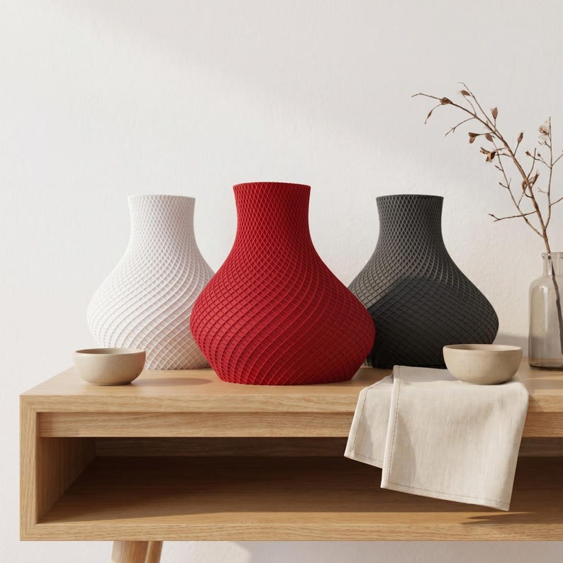 May include: Three decorative vases in white, red, and gray, with a textured, woven design. The vases are displayed on a light wood table, alongside small bowls and a folded linen cloth. A minimalist home decor theme.
