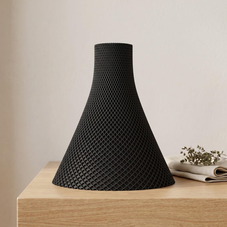 Peut inclure: Un vase noir en forme de c&ocirc;ne avec un motif textur&eacute; en forme de treillis. Le vase a une base large qui se r&eacute;tr&eacute;cit vers le haut jusqu'&agrave; un col plus &eacute;troit. Il repose sur une surface en bois de couleur claire. Un tissu pli&eacute; et une brindille de fleurs s&eacute;ch&eacute;es sont en arri&egrave;re-plan.