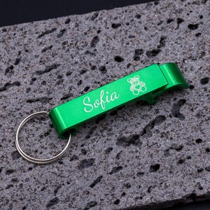 Grüner Flaschenöffner Schlüsselanhänger aus Aluminium mit Gravur „Sofia“ und Bärchen-Symbol. Praktischer Kapselheber mit Schlüsselring, stabil, leicht und personalisierbar. Ideal als Geschenk oder individuelles Accessoire für den Schlüsselbund, kombiniert Funktionalität und persönliche Note. Präsentiert auf grauem Steinuntergrund für stilvolle Produktdarstellung.