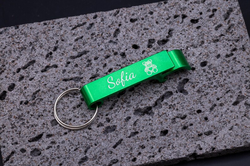 Grüner Flaschenöffner Schlüsselanhänger aus Aluminium mit Gravur „Sofia“ und Bärchen-Symbol. Praktischer Kapselheber mit Schlüsselring, stabil, leicht und personalisierbar. Ideal als Geschenk oder individuelles Accessoire für den Schlüsselbund, kombiniert Funktionalität und persönliche Note. Präsentiert auf grauem Steinuntergrund für stilvolle Produktdarstellung.