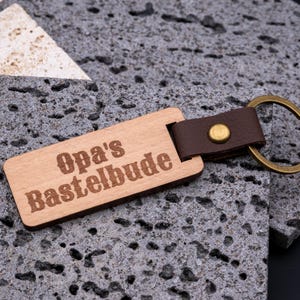 Rustikaler Schlüsselanhänger aus Holz mit Gravur „Opa’s Bastelbude“. Der rechteckige Holzanhänger ist mit braunem Kunstlederband und stabilem Messingring versehen, wodurch er robust und langlebig wird. Die humorvolle Gravur macht den Anhänger zu einem originellen Geschenk für Großväter, Heimwerker oder Bastler. Kombination aus natürlicher Holzoptik, edlem Lederlook und praktischer Funktion für Schlüsselbund oder Werkstatt.