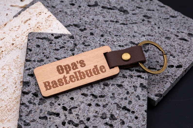 Rustikaler Schlüsselanhänger aus Holz mit Gravur „Opa’s Bastelbude“. Der rechteckige Holzanhänger ist mit braunem Kunstlederband und stabilem Messingring versehen, wodurch er robust und langlebig wird. Die humorvolle Gravur macht den Anhänger zu einem originellen Geschenk für Großväter, Heimwerker oder Bastler. Kombination aus natürlicher Holzoptik, edlem Lederlook und praktischer Funktion für Schlüsselbund oder Werkstatt.