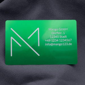 Grüne Visitenkarte aus Metall mit Firmenlogo, Anschrift, Telefonnummer und E-Mail. Das klare, weiße Schriftbild hebt sich gut von der glänzenden Aluminiumoberfläche ab. Die robuste Karte eignet sich hervorragend für den geschäftlichen Einsatz und vermittelt Professionalität, Exklusivität und Beständigkeit, perfekt für Unternehmer, die mit individuellen Visitenkarten auffallen möchten.