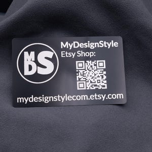Schwarze Aluminium-Visitenkarte mit MDS-Logo, Etsy-Shop-Adresse und QR-Code, auf Stoff fotografiert. Die Gravur ist klar und langlebig, die Karte im Kreditkartenformat wirkt edel und stabil. Ideal für Branding, Kundenbindung und kreative Businessideen.