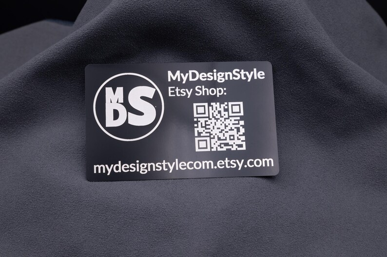 Schwarze Aluminium-Visitenkarte mit MDS-Logo, Etsy-Shop-Adresse und QR-Code, auf Stoff fotografiert. Die Gravur ist klar und langlebig, die Karte im Kreditkartenformat wirkt edel und stabil. Ideal für Branding, Kundenbindung und kreative Businessideen.