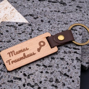 Personalisierter Schlüsselanhänger aus Holz mit Gravur „Mamas Traumhaus“ und kleinem Schlüssel-Symbol. Der rechteckige Holzanhänger ist mit einem braunen Kunstlederband und stabilem Messingring versehen, was ihm eine edle, langlebige Optik verleiht. Ein originelles Geschenk für Frauen und Mütter, ideal zur Einweihung, für den neuen Hausschlüssel oder als persönliches Accessoire. Kombination aus Holzdesign und Lederoptik.