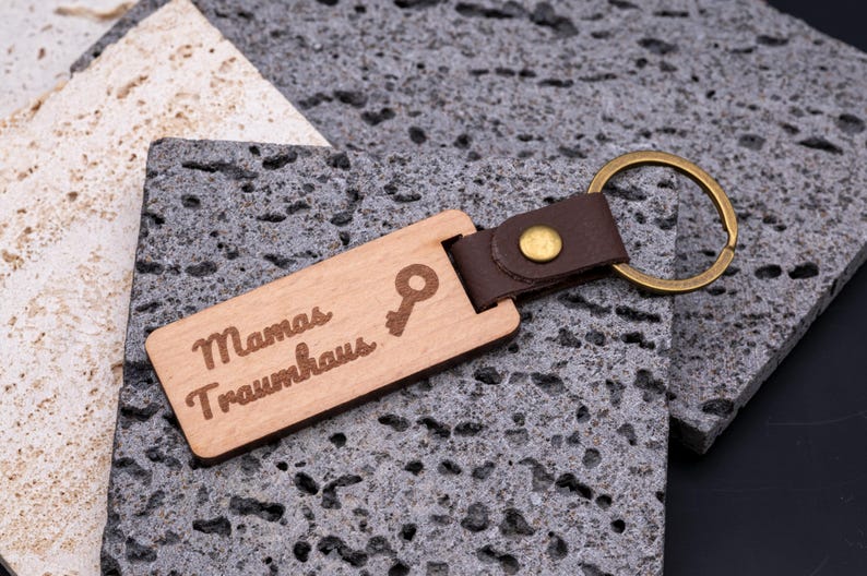 Personalisierter Schlüsselanhänger aus Holz mit Gravur „Mamas Traumhaus“ und kleinem Schlüssel-Symbol. Der rechteckige Holzanhänger ist mit einem braunen Kunstlederband und stabilem Messingring versehen, was ihm eine edle, langlebige Optik verleiht. Ein originelles Geschenk für Frauen und Mütter, ideal zur Einweihung, für den neuen Hausschlüssel oder als persönliches Accessoire. Kombination aus Holzdesign und Lederoptik.