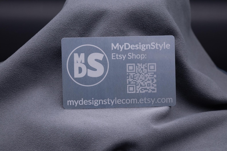 Visitenkarte aus Aluminium in Silber mit matter Oberfläche und Gravur des Logos „MDS“, dem Hinweis auf den Etsy-Shop sowie einem QR-Code. Die edle Metallkarte kombiniert modernes Design mit Funktionalität und ist perfekt geeignet für Unternehmen, Designer und Kreative. Eine langlebige und stilvolle Alternative zur klassischen Papierkarte, ideal für Branding, Networking und Marketingaktionen.