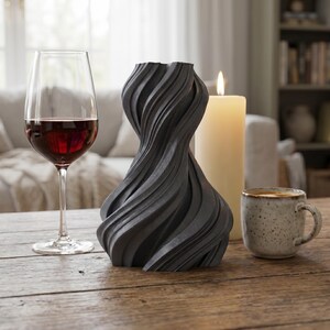 K&ouml;nnte beinhalten: Eine dunkelgraue, gedrehte Vase mit strukturierter Oberfl&auml;che steht auf einem Holztisch. Ein Glas Rotwein, eine brennende Kerze und eine gesprenkelte Tasse stehen ebenfalls auf dem Tisch. Die Vase hat ein einzigartiges, wirbelndes Design.