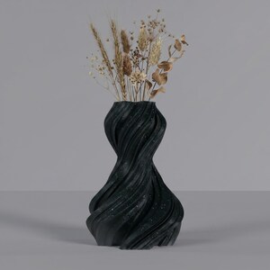 K&ouml;nnte beinhalten: Eine schwarze, strukturierte Vase mit einem Wirbeldesign, &uuml;bers&auml;t mit wei&szlig;en Flecken. Die Vase enth&auml;lt getrockneten Weizen, Gr&auml;ser und Eukalyptuszweige. Die Vase hat eine einzigartige, k&uuml;nstlerische Form und ist ein dekoratives Wohnaccessoire.