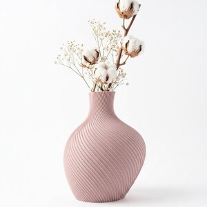 Puede incluir: Un jarr&oacute;n de color rosa polvoriento con un dise&ntilde;o texturizado y arremolinado contiene tallos de algod&oacute;n y delicadas flores blancas. El jarr&oacute;n tiene una base redondeada y un cuello delgado, creando una est&eacute;tica elegante.