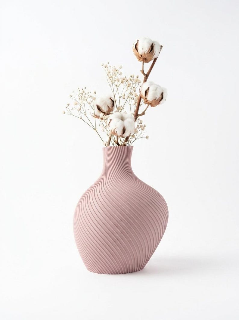Puede incluir: Un jarr&oacute;n de color rosa polvoriento con un dise&ntilde;o texturizado y arremolinado contiene tallos de algod&oacute;n y delicadas flores blancas. El jarr&oacute;n tiene una base redondeada y un cuello delgado, creando una est&eacute;tica elegante.