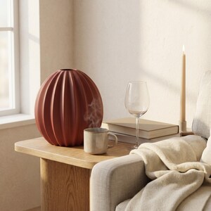 May include: A terracotta-coloured, ribbed vase, a small mug, a wine glass, and books on a wooden side table. A lit candle and a blanket draped over a sofa arm create a cosy atmosphere. The vase is a prominent feature.