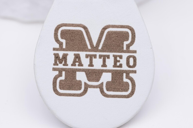 Detailaufnahme des Namens „Matteo“, kunstvoll graviert in goldbrauner Farbe auf weißem veganem Kunstleder. Das Monogramm-Design sorgt für einen eleganten und modernen Look, während die Lasergravur durch Präzision und Klarheit besticht. Der Schlüsselanhänger ist langlebig, angenehm in der Hand und ein ideales personalisiertes Geschenk für Geburtstage, Valentinstag oder als stilvolles Accessoire im Alltag.