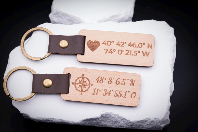 Zwei personalisierte Holz Schlüsselanhänger nebeneinander auf weißem Stein, Gravuren mit Herzsymbol und Koordinaten sowie Kompasssymbol mit Zahlen. Beide Anhänger bestehen aus hellem Holz, sind mit braunem Lederband und stabilem Metallring verbunden. Ideal als Geschenkidee für Paare, Reisende oder als Andenken an Lieblingsorte. Die Gravuren verleihen dem Schlüsselanhänger eine ganz persönliche Bedeutung und machen ihn einzigartig.