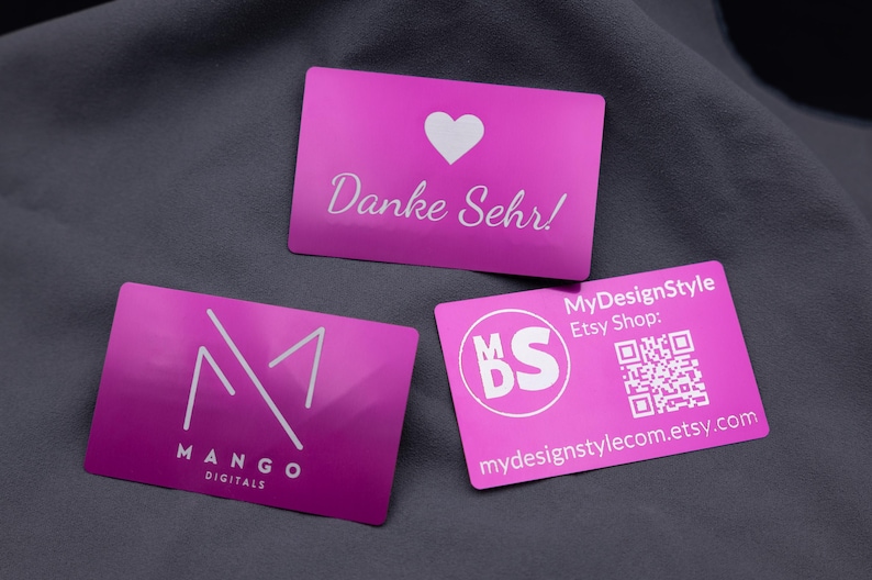 Edle lila Visitenkarten aus robustem Aluminium mit Gravur. Das Set zeigt drei verschiedene Designs: eine Karte mit Herzsymbol und „Danke Sehr“-Gravur, eine mit dem modernen Mango-Logo sowie eine MyDesignStyle-Karte mit QR-Code und Shop-Adresse. Die metallisch glänzende Oberfläche in Lila wirkt elegant und auffällig, ideal für Business, Branding oder persönliche Danksagungen.