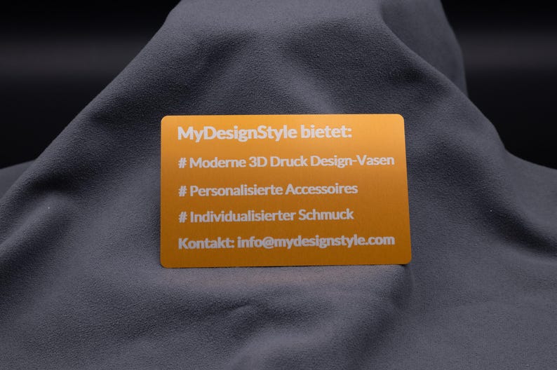 Hellgoldene Aluminiumkarte mit Gravur der MyDesignStyle-Angebote: moderne 3D-Druck Vasen, personalisierte Accessoires und individualisierter Schmuck. Zusätzlich mit Kontakt-E-Mail versehen. Die gravierte Metallkarte vereint Werbewirkung mit hochwertigem Design und eignet sich perfekt als elegante Informationskarte, Beileger oder Präsentationskarte für Online-Shops.