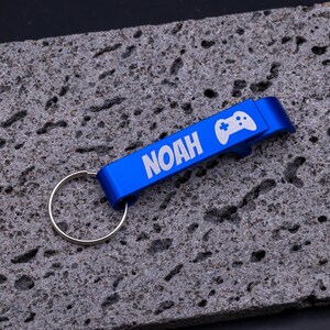 Blauer Flaschenöffner Schlüsselanhänger aus Aluminium mit Gravur „NOAH“ und Symbol eines Game-Controllers. Praktischer Kapselheber mit Schlüsselring, robust und leicht, ideal als personalisiertes Geschenk für Gamer, Freunde oder Familie. Individuell gravierbar mit Namen, Symbolen oder Logos, modernes Accessoire für Schlüsselbund und Flaschenöffnen, dargestellt auf grauem Steinuntergrund.