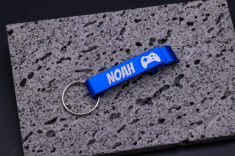 Blauer Flaschenöffner Schlüsselanhänger aus Aluminium mit Gravur „NOAH“ und Symbol eines Game-Controllers. Praktischer Kapselheber mit Schlüsselring, robust und leicht, ideal als personalisiertes Geschenk für Gamer, Freunde oder Familie. Individuell gravierbar mit Namen, Symbolen oder Logos, modernes Accessoire für Schlüsselbund und Flaschenöffnen, dargestellt auf grauem Steinuntergrund.