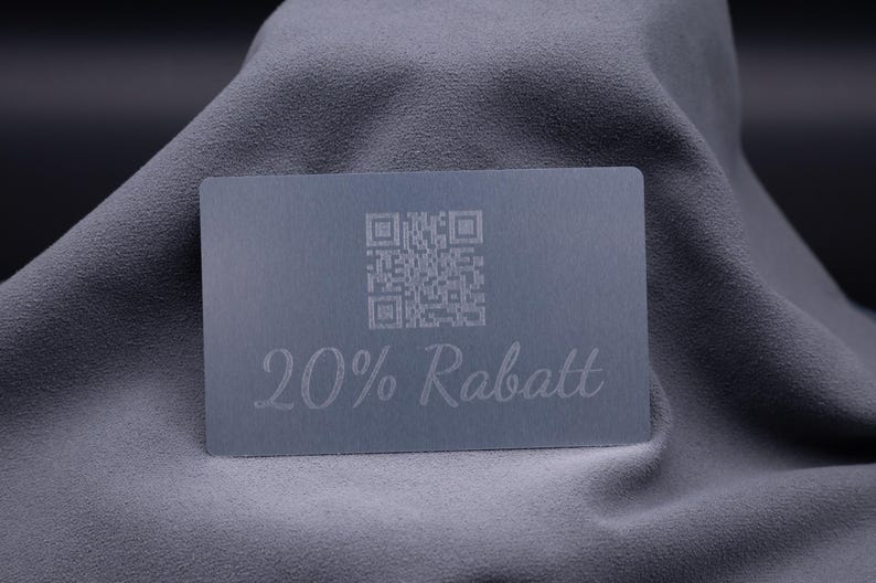 Metallische Rabattkarte aus Aluminium in Silber mit Gravur „20% Rabatt“ sowie QR-Code, auf grauem Stoff präsentiert. Die Karte kombiniert modernes Design mit praktischer Funktionalität, wirkt edel und ist robust im Einsatz. Ideal als exklusive Werbekarte, Gutscheinkarte oder Marketinginstrument für Unternehmen, die ihre Kunden mit einem eleganten und langlebigen Medium begeistern möchten.