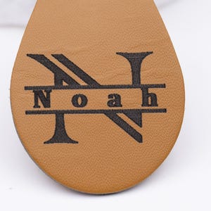 Nahaufnahme der Gravur eines personalisierten Schlüsselanhängers aus hellbraunem veganem Kunstleder. Zu sehen ist das elegante Monogramm-Design „Noah“ mit großem Buchstaben „N“ und feiner Lasergravur in Schwarz. Die Textur des Leders wirkt weich und hochwertig, ideal für individuelle Geschenke mit Namen oder Initialen.