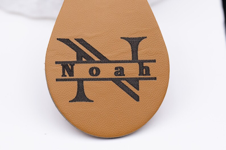 Nahaufnahme der Gravur eines personalisierten Schlüsselanhängers aus hellbraunem veganem Kunstleder. Zu sehen ist das elegante Monogramm-Design „Noah“ mit großem Buchstaben „N“ und feiner Lasergravur in Schwarz. Die Textur des Leders wirkt weich und hochwertig, ideal für individuelle Geschenke mit Namen oder Initialen.
