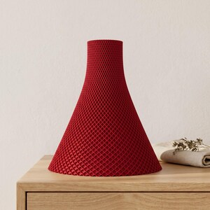 Peut inclure: Un vase rouge en forme de c&ocirc;ne avec une surface textur&eacute;e en forme de treillis. Le vase a une base large qui se r&eacute;tr&eacute;cit vers le haut. Le vase est fait d'un mat&eacute;riau solide et repose sur une surface en bois.