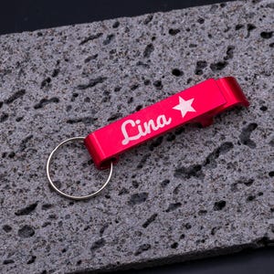 Roter Flaschenöffner Schlüsselanhänger aus Aluminium mit Gravur „Lina“ und Stern-Symbol. Der leichte und robuste Kapselheber besitzt einen Schlüsselring und ist personalisierbar, ideal als Geschenk oder praktisches Accessoire für den Alltag. Edle Gravur verleiht dem Schlüsselanhänger eine individuelle Note. Präsentiert auf grauem Steinuntergrund für eine moderne Produktdarstellung.