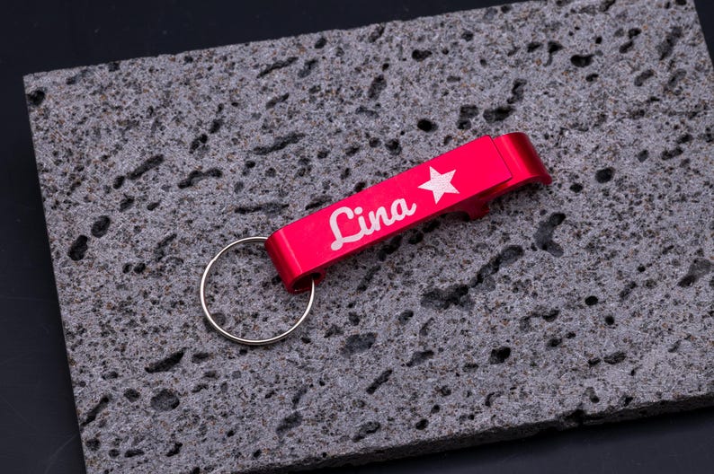 Roter Flaschenöffner Schlüsselanhänger aus Aluminium mit Gravur „Lina“ und Stern-Symbol. Der leichte und robuste Kapselheber besitzt einen Schlüsselring und ist personalisierbar, ideal als Geschenk oder praktisches Accessoire für den Alltag. Edle Gravur verleiht dem Schlüsselanhänger eine individuelle Note. Präsentiert auf grauem Steinuntergrund für eine moderne Produktdarstellung.