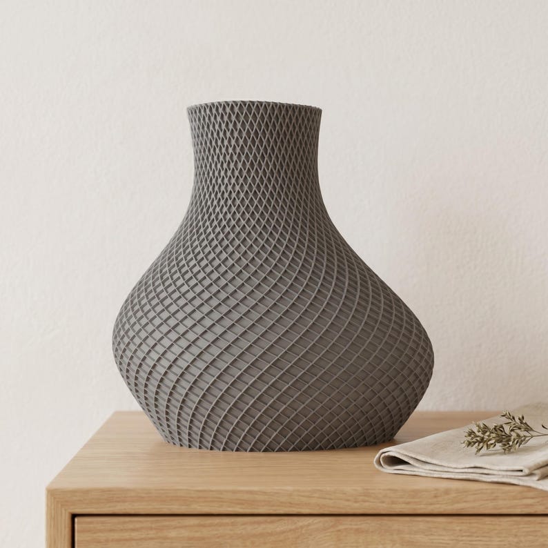 May include: A gray decorative vase with a unique, textured, lattice-like pattern. The vase has a wide, rounded base that tapers to a narrower neck and opening. It sits on a wooden surface with a folded cloth and sprig of greenery.