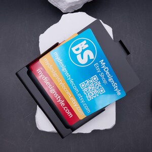 Offenes schwarzes Metall-Visitenkartenetui mit drei farbigen Karten (rot, gelb, blau). Auf weißem Stein im Studio fotografiert. Das Etui ist stabil, leicht und personalisierbar durch Lasergravur. Ideal als Geschenk für Männer und Frauen im Business-Umfeld, die Visitenkarten stilvoll präsentieren möchten. Perfekt für Büro, Networking und Kundenmeetings.